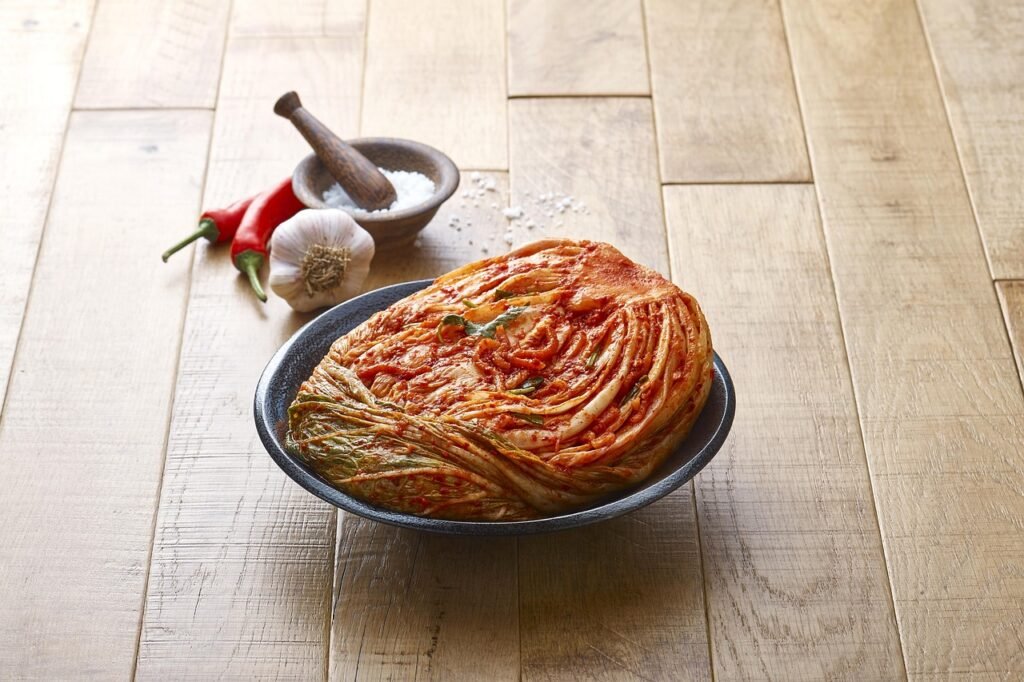 Whole head of red napa cabbage kimchi in a dark bowl on a wooden table beside garlic and chilies
