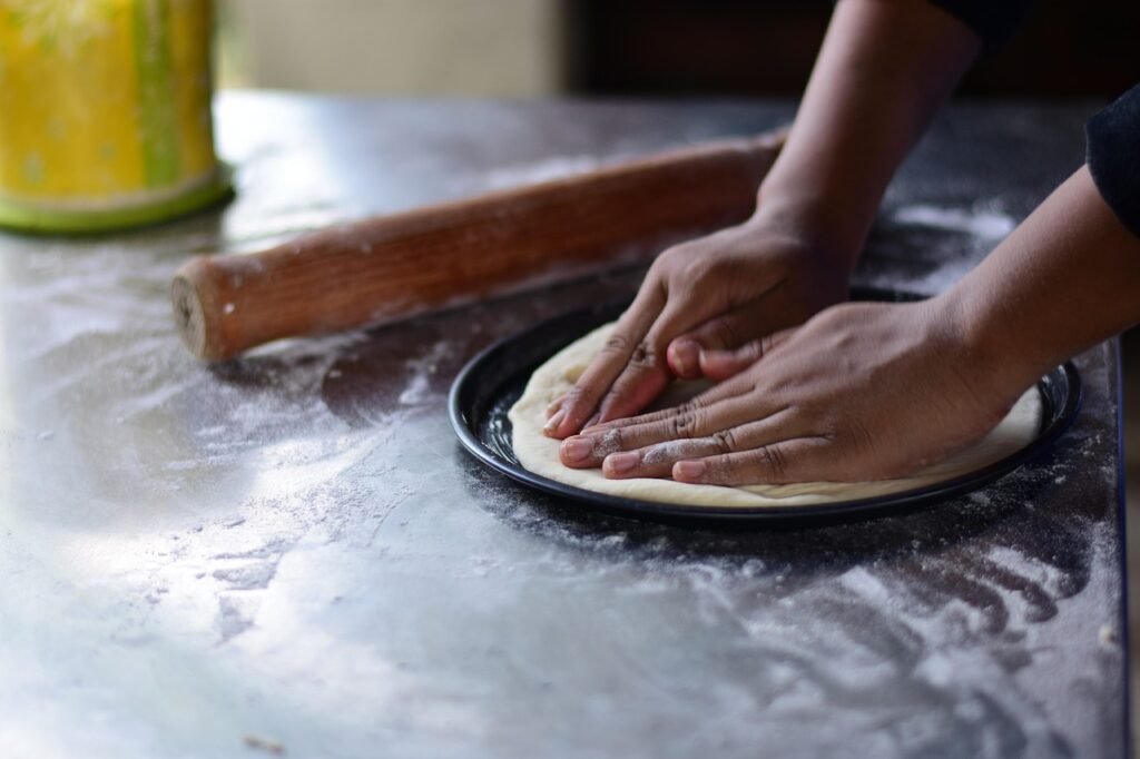 Hands pressing pizza dough flat into a round dark pan on a floured counter