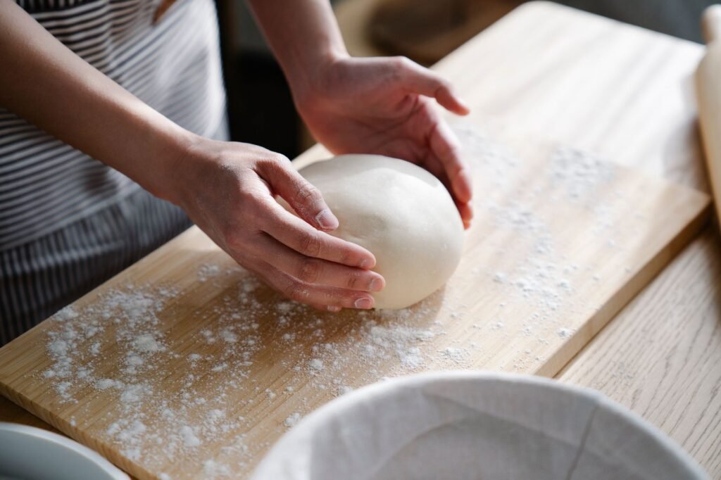 Hands shaping a smooth ball of pizza dough on a floured wooden board