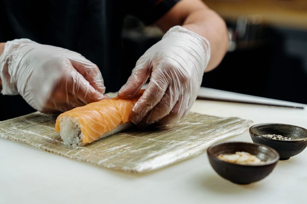 Gloved hands rolling a salmon sushi roll on a bamboo mat beside small dipping bowls