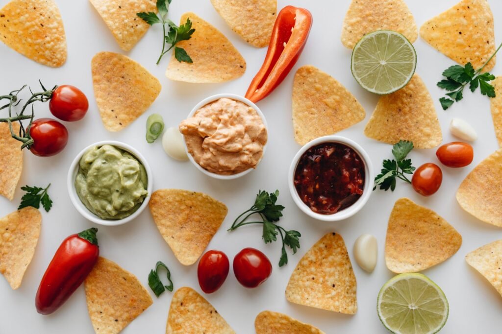 Overhead spread of tortilla chips with guacamole, salsa, limes, tomatoes and red peppers on white