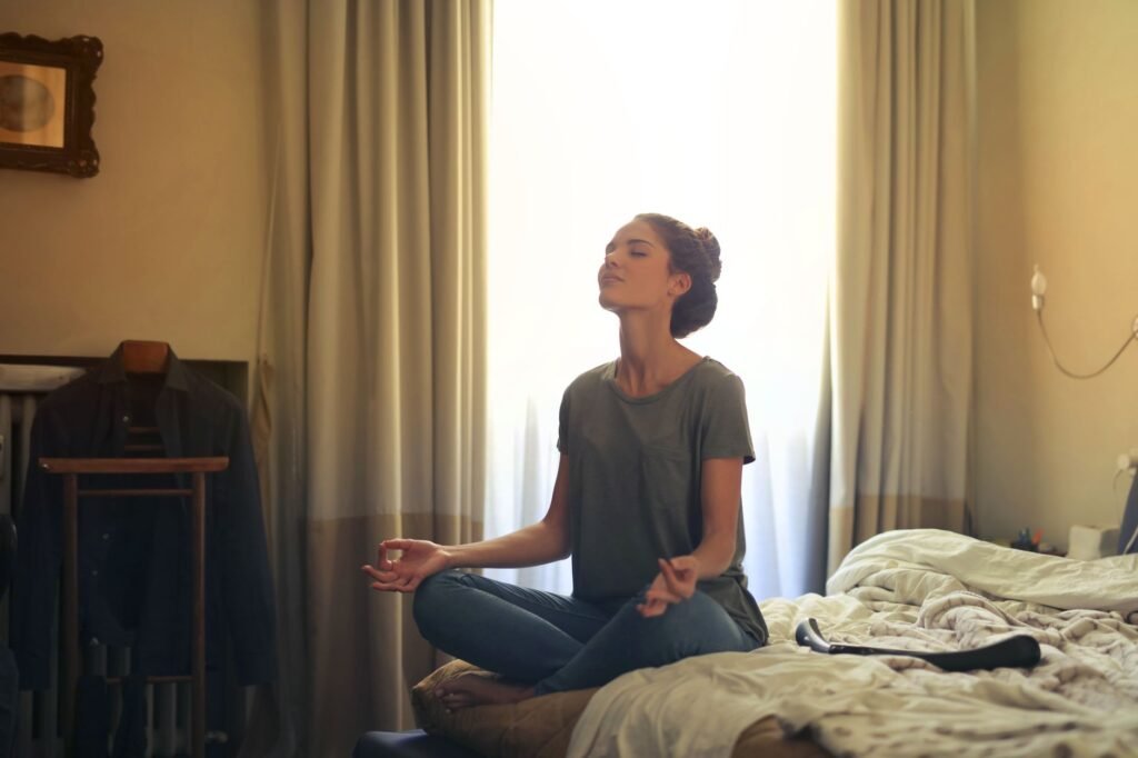 Woman meditating cross legged on a bed with eyes closed in a sunlit room