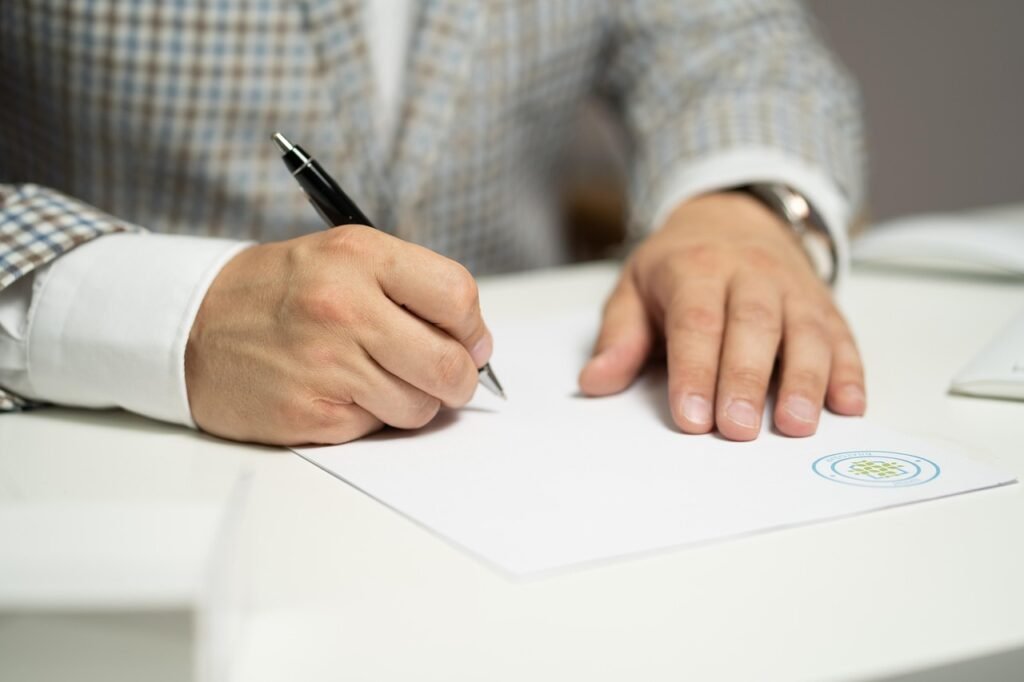 Man in a checked jacket signing a paper document with a black pen at a table