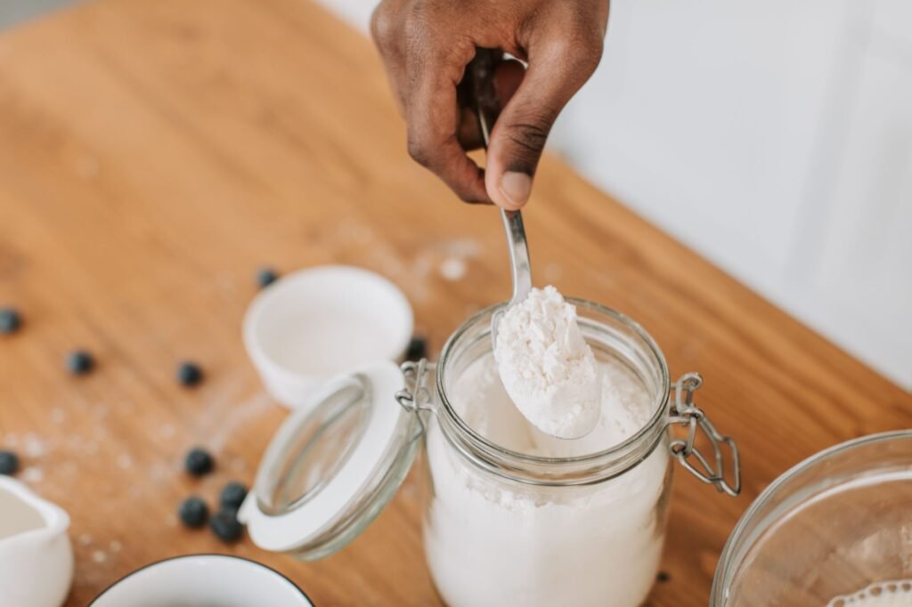 Hand scooping flour with a spoon from a clip top glass jar on a wooden table