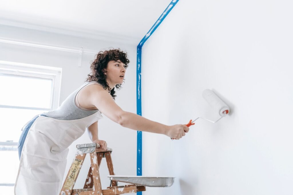 Woman on a stepladder rolling white paint onto a wall edged with blue painter's tape