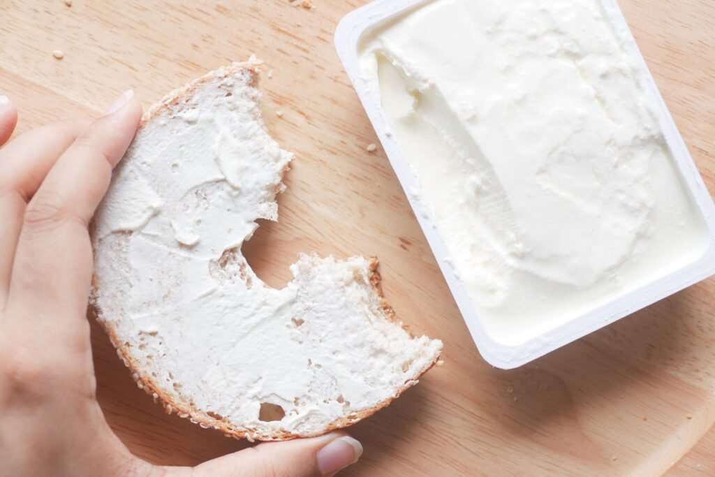 Close-up of a hand holding a bagel with creamy cheese spread next to an open container
