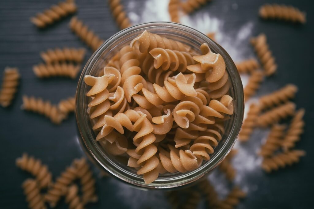 Open glass jar filled with dry spiral pasta surrounded by loose pasta on a dark surface
