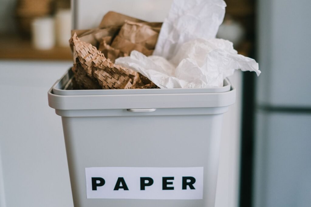 Gray kitchen bin labeled paper overflowing with crumpled brown and white paper