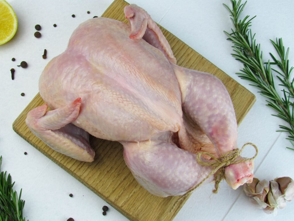 Raw whole chicken on a cutting board with rosemary, garlic, and peppercorns
