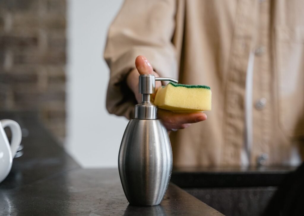 Hand holding a yellow and green kitchen sponge above a steel soap dispenser on a counter