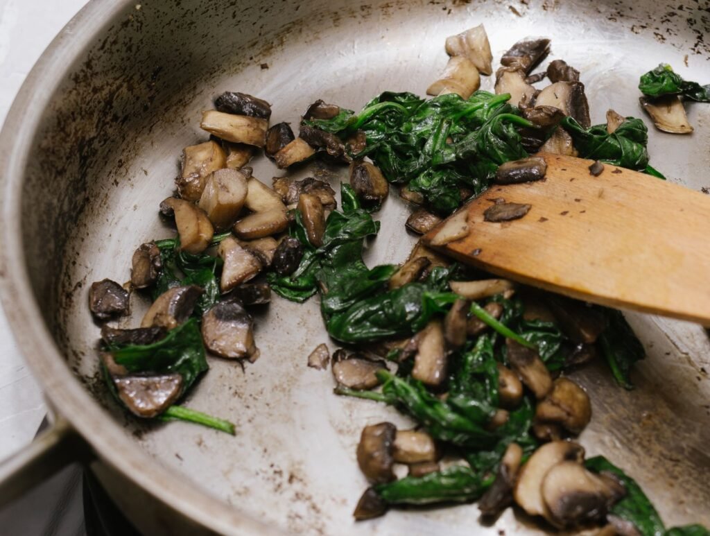 Wilted spinach and sliced mushrooms cooking in a pan with a wooden spatula