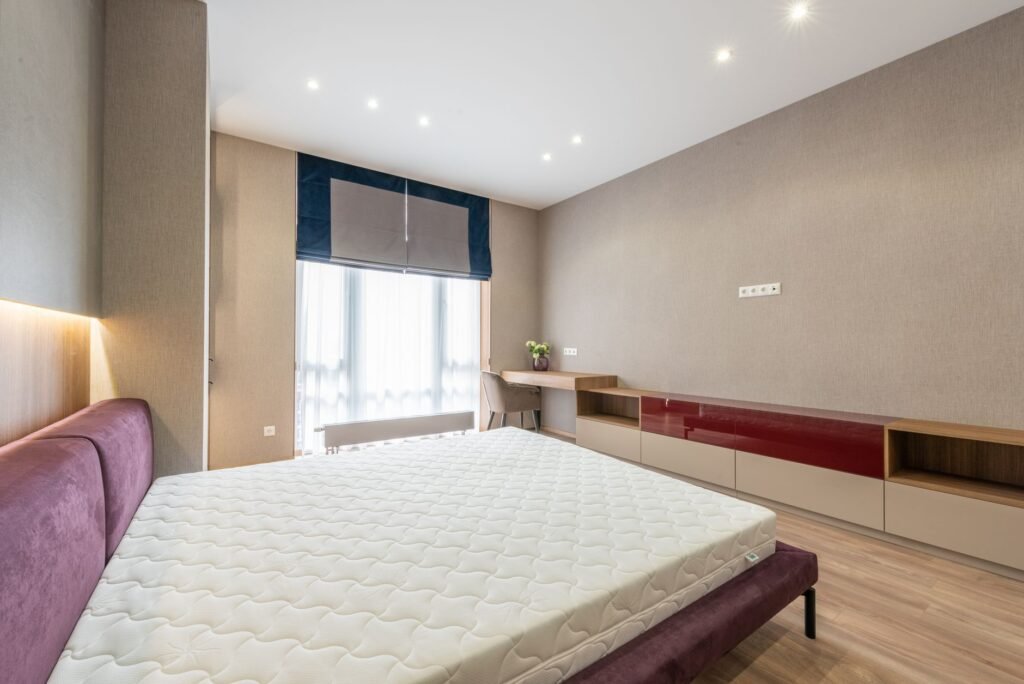 Modern bedroom with white mattress on bed placed against wooden cabinet and table with flowerpot at wall wi...
