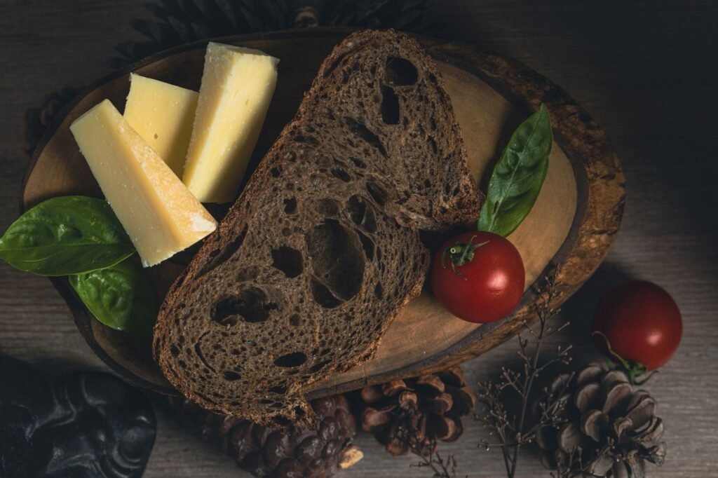 A dark rustic bread with cheese wedges, cherry tomatoes, and basil on a wooden board
