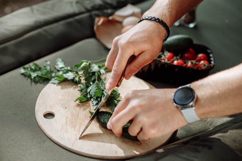 Hands cutting cucumbers and parsley with cherry tomatoes nearby on a wooden board.