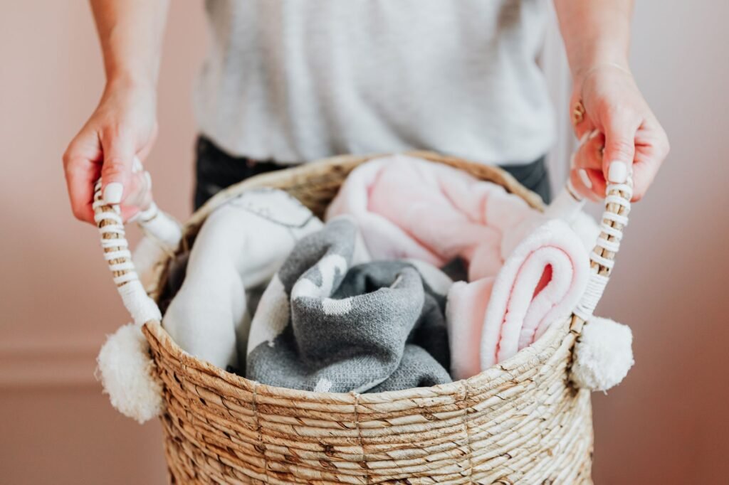 Person carrying a woven basket filled with soft folded blankets in pink, gray and white