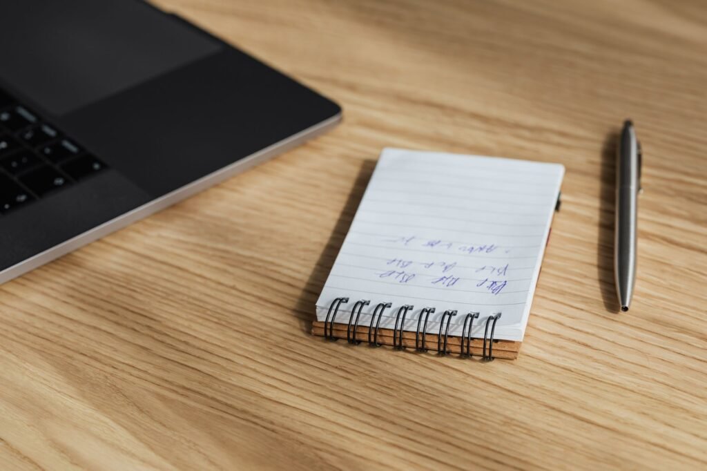 Small spiral notepad with handwritten lines and a silver pen on a wooden desk beside a laptop