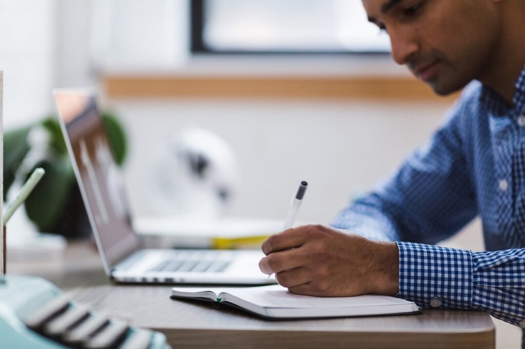 Man in a checked shirt writing in a notebook beside an open laptop at a desk