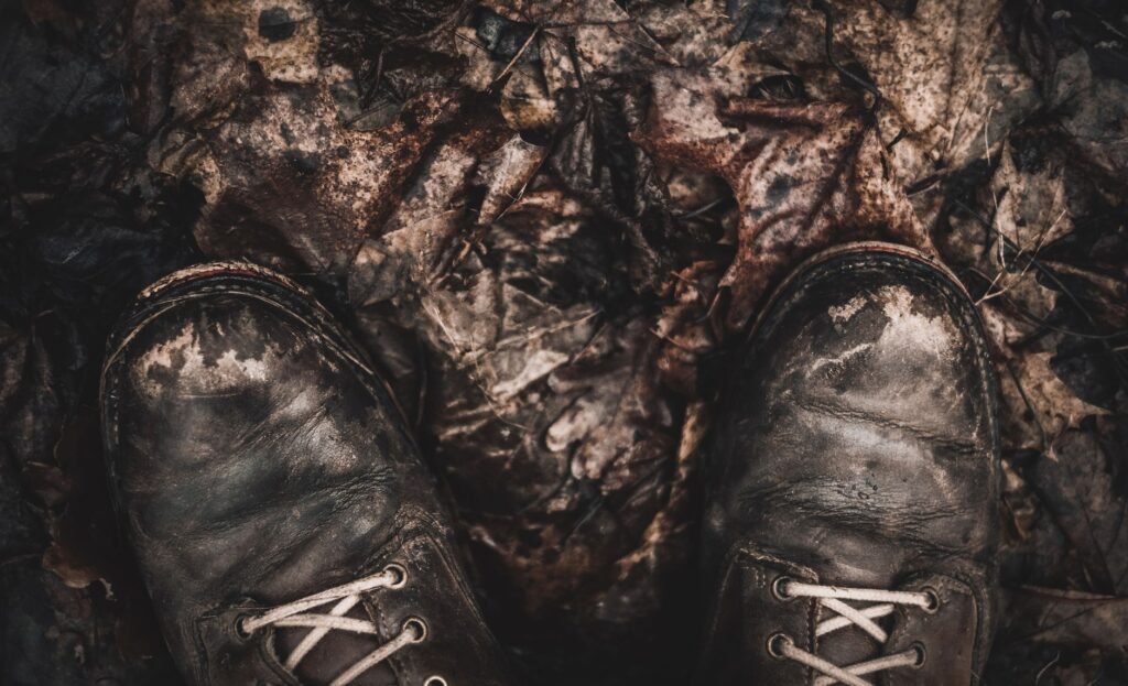 A pair of heavily worn dark leather lace up boots seen from above on muddy ground