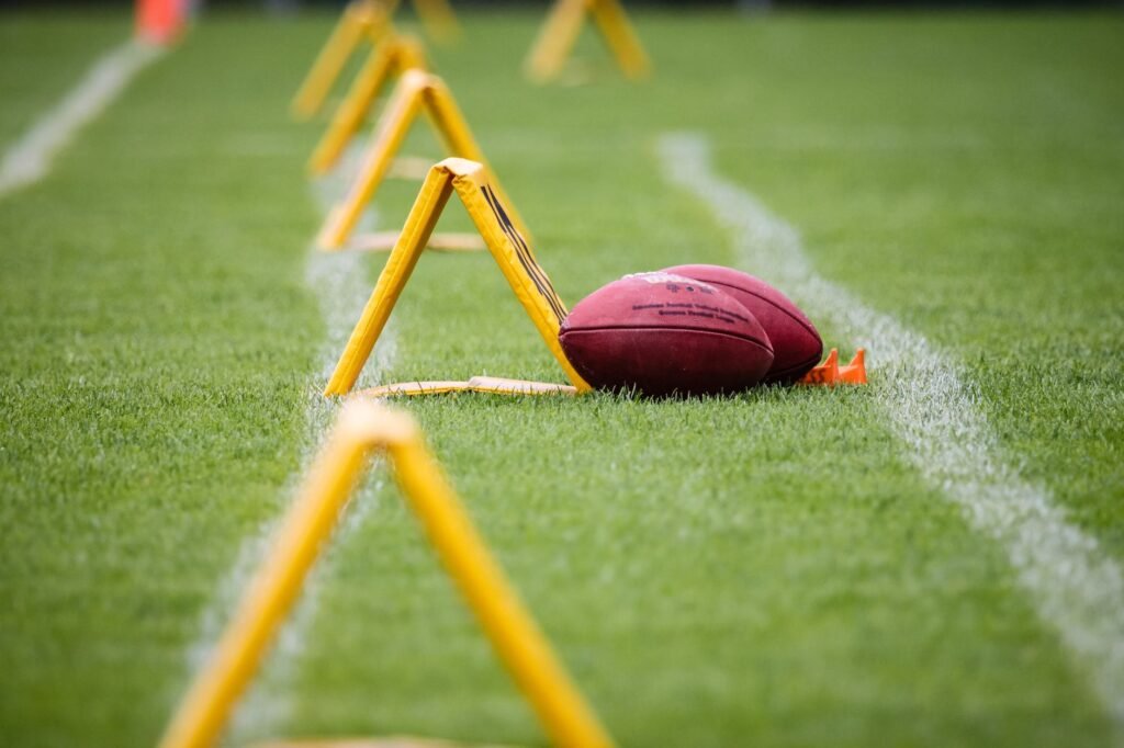 A brown leather American football resting on green turf beside yellow training hurdles