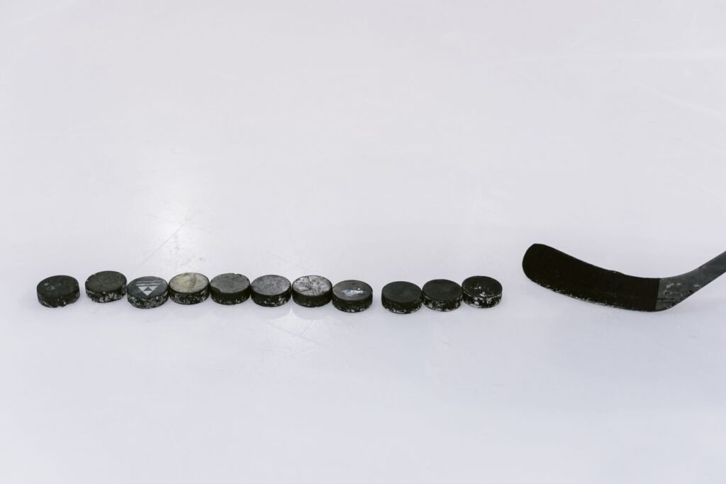 A row of black hockey pucks lined up on white ice beside the blade of a hockey stick