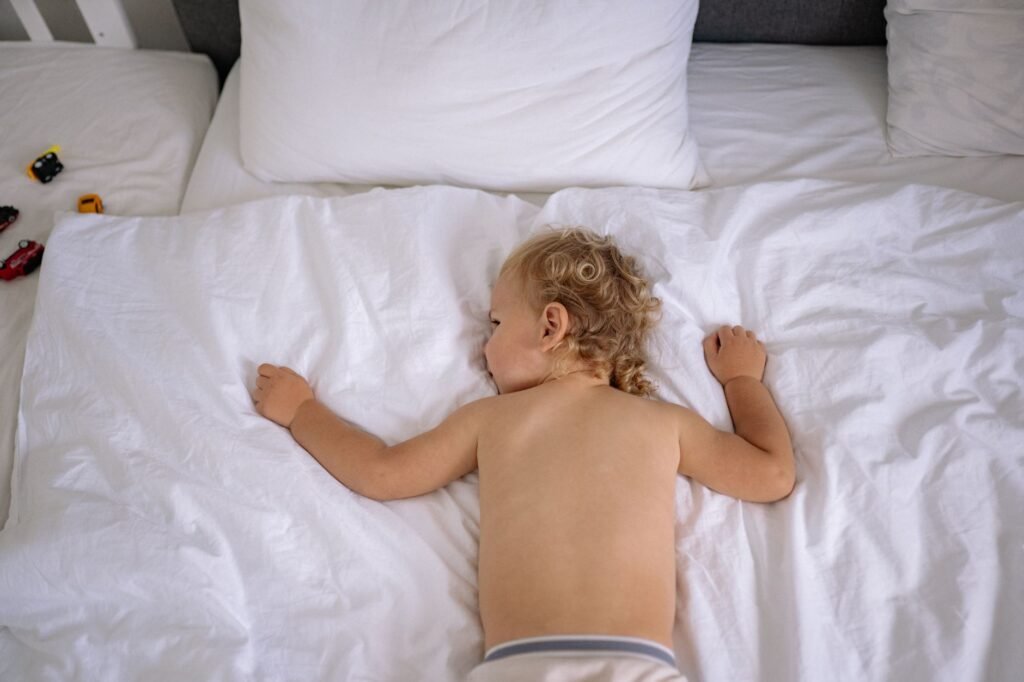 A toddler sleeps comfortably on a white bed, surrounded by toys, capturing a serene indoor scene.