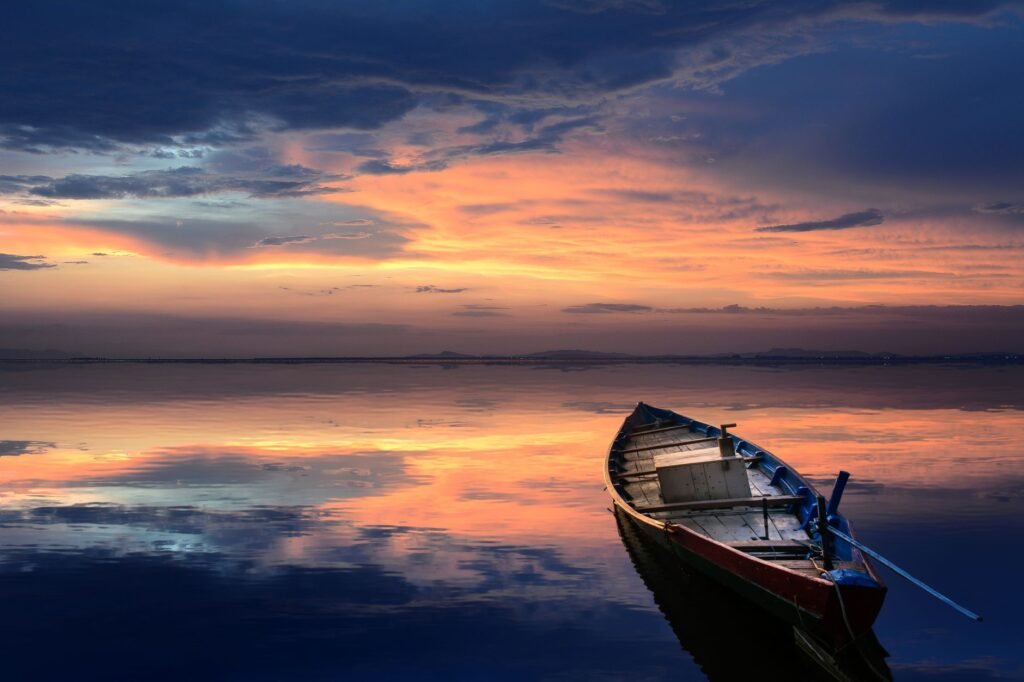 A serene scene of a wooden boat floating on a calm sea at sunset, reflecting vibrant colors and a tranquil ...