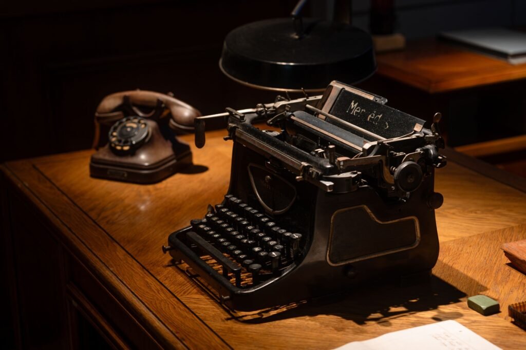 An antique black manual typewriter on a polished wooden desk beside a rotary telephone