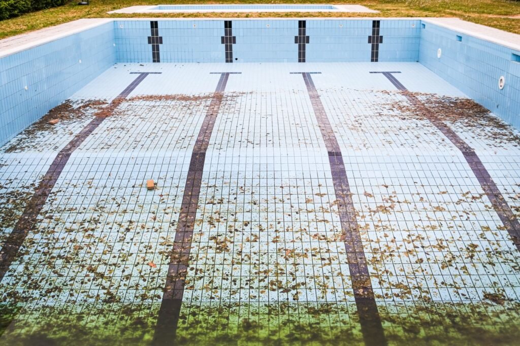 A drained outdoor swimming pool with pale blue tiles and fallen leaves across the floor