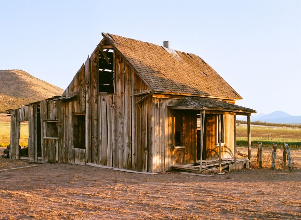 A rustic abandoned cabin sits in a sunlit desert landscape, capturing a timeless rural charm