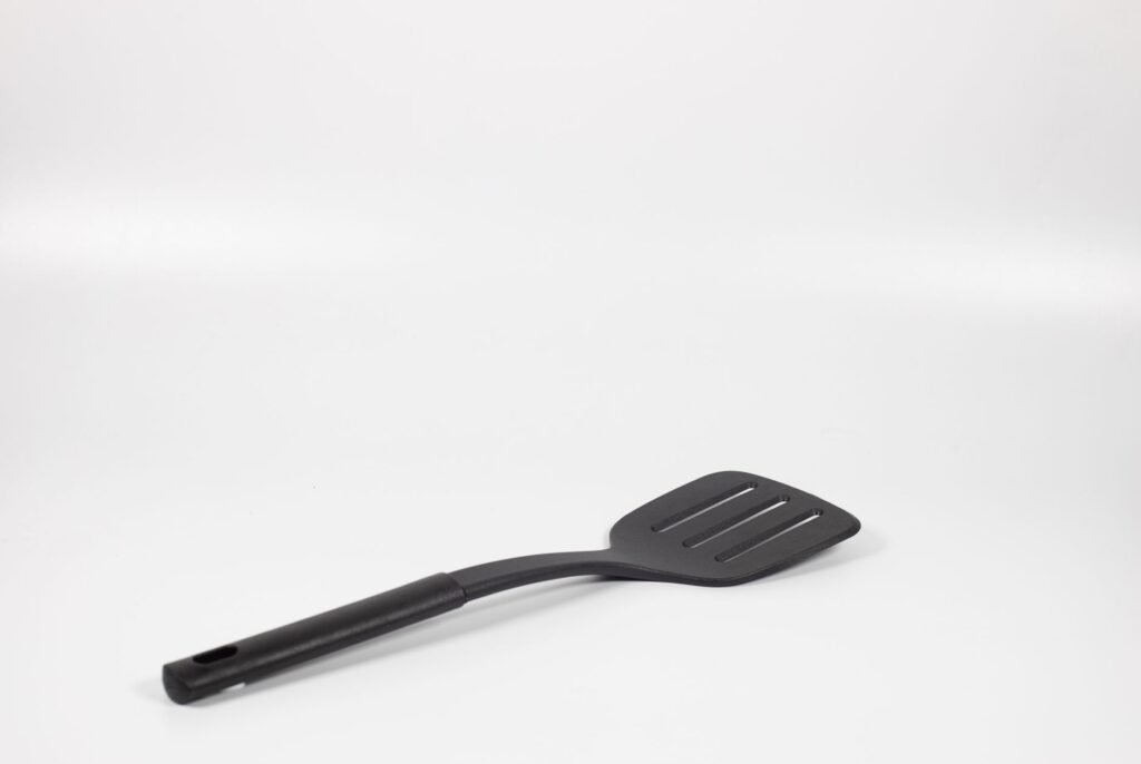 Black slotted spatula lying on a plain white surface with soft shadows