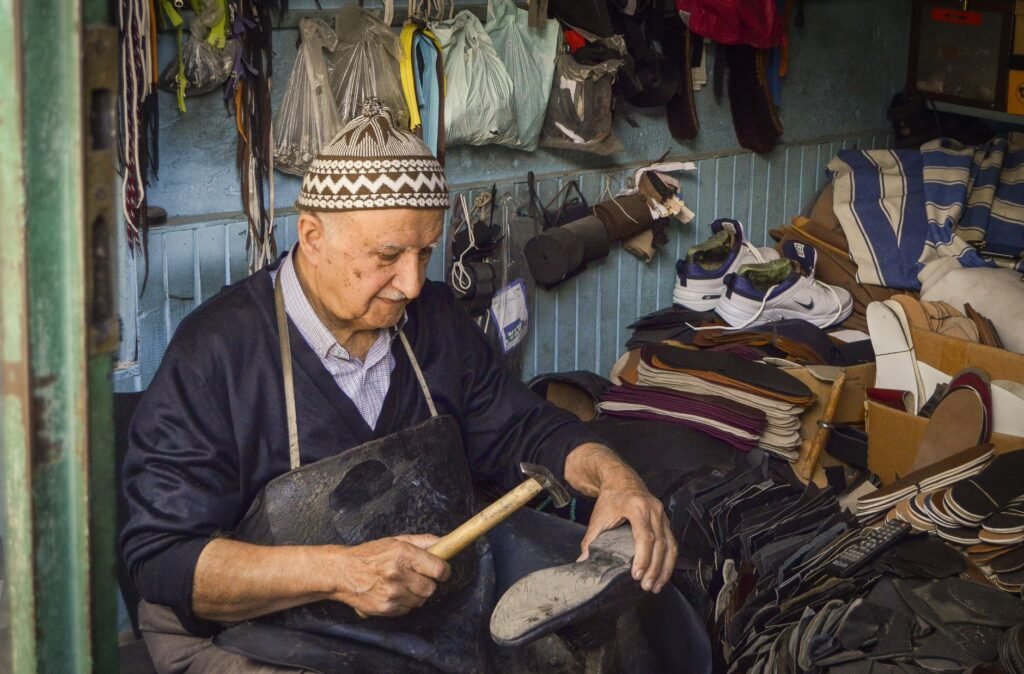 Older cobbler in a knit cap hammering the sole of a shoe in a cluttered workshop