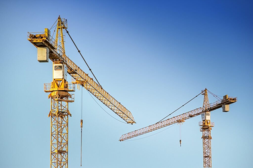 Two yellow tower cranes with long lattice arms reaching across a clear blue sky
