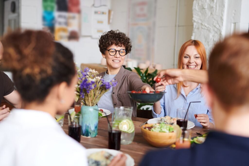 Friends smiling and passing dishes across a dinner table set with flowers, salad, and drinks