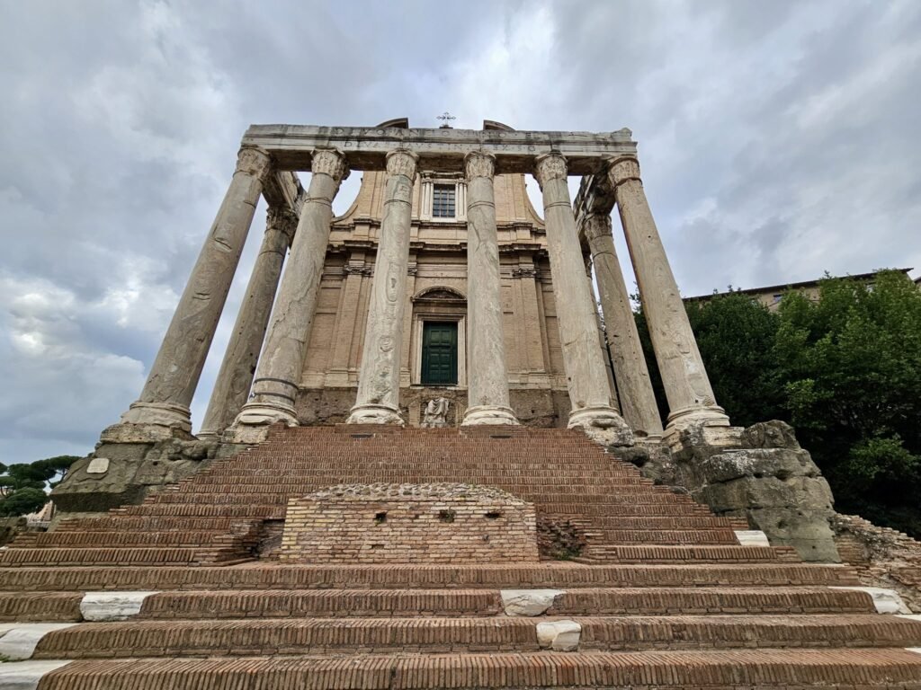 The grand front view of the Temple of Antoninus and Faustina, with tall marble columns and brick steps lead...