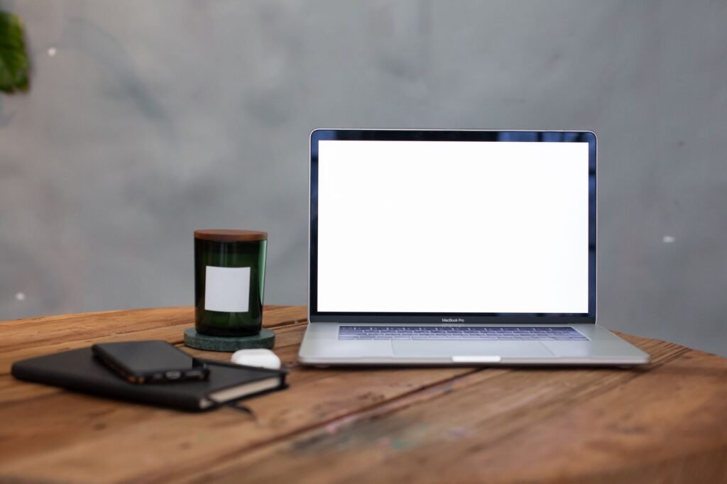 Open laptop with a blank white screen on a wooden table beside a notebook and candle