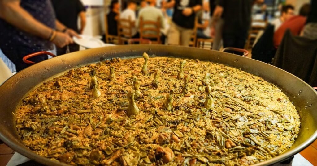 A huge pan of Valencian paella with green beans and chicken drumsticks on a table surrounded by people, some of whom are