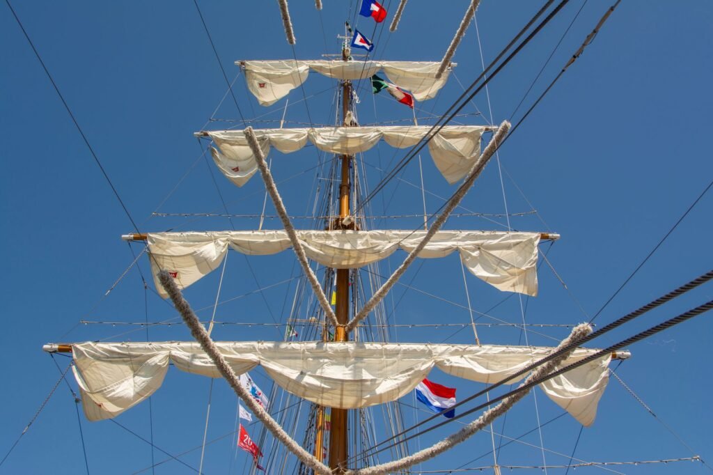 Close-up of a sailing ship's mast with white sails and colorful flags against a bright blue sky