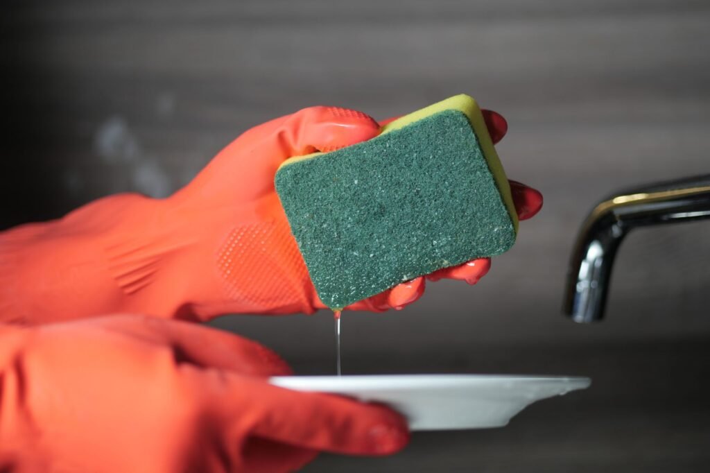 Gloved hand squeezing water from a green and yellow kitchen sponge over a plate