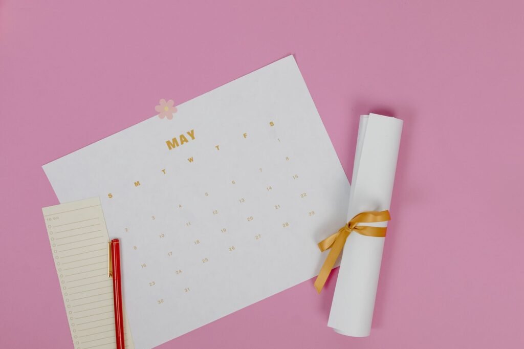 Printed monthly calendar sheet on a pink surface beside a rolled paper tied with gold ribbon