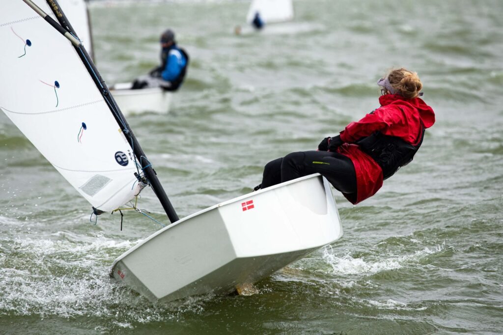 Sailor in a red jacket leaning out as a small white sailboat tilts in choppy water