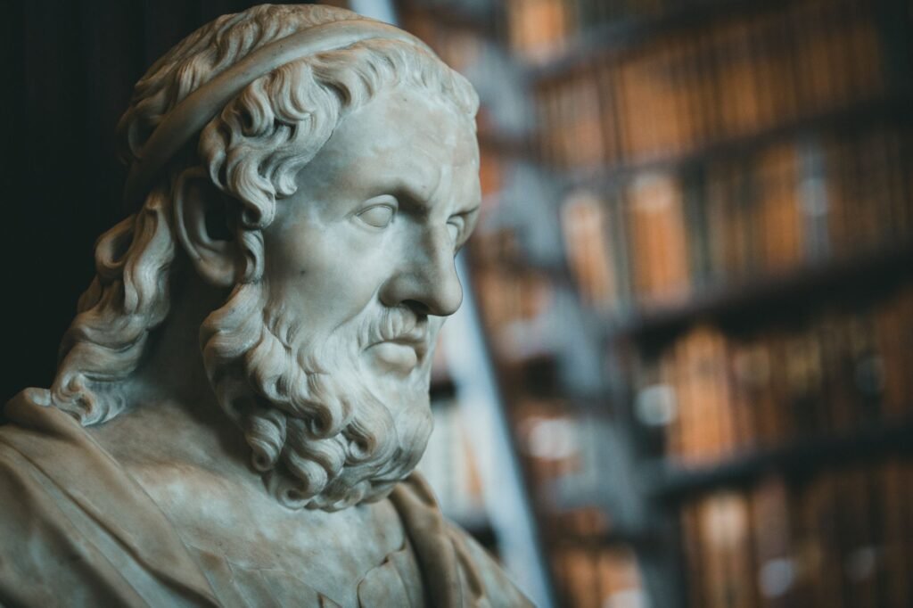 Stone bust of a bearded man with curly hair in front of blurred library shelves