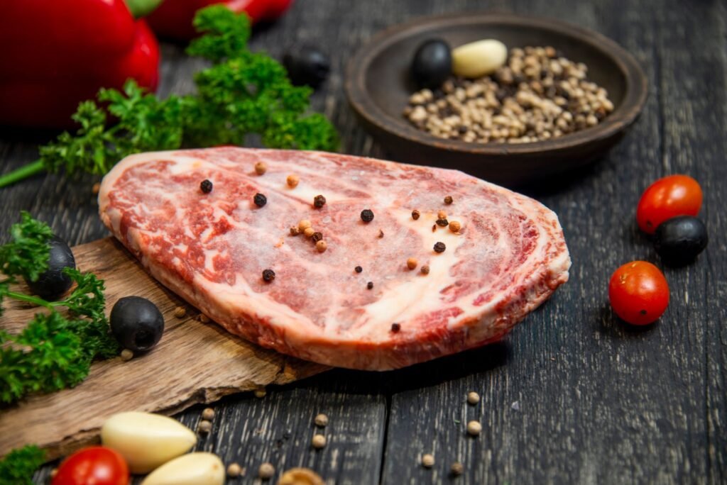 Raw marbled beef steak on a wooden board surrounded by peppercorns, cherry tomatoes, and fresh herbs