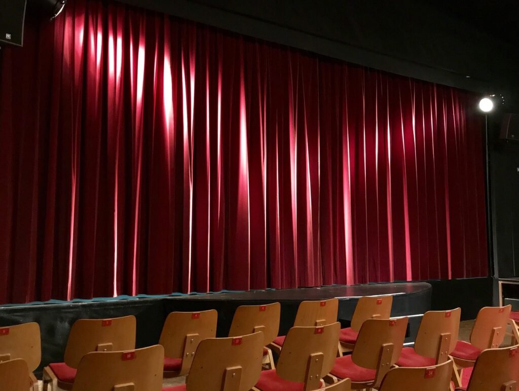 Red velvet stage curtain lit by spotlights above rows of empty audience chairs
