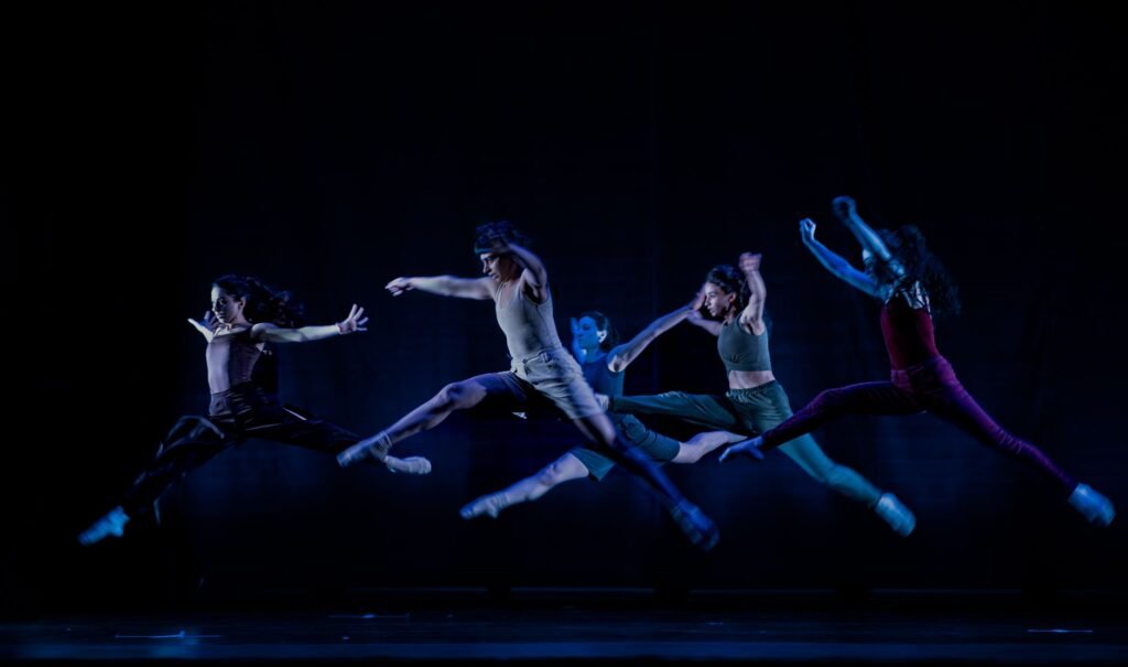 Four dancers leaping in midair across a dark stage lit with blue light