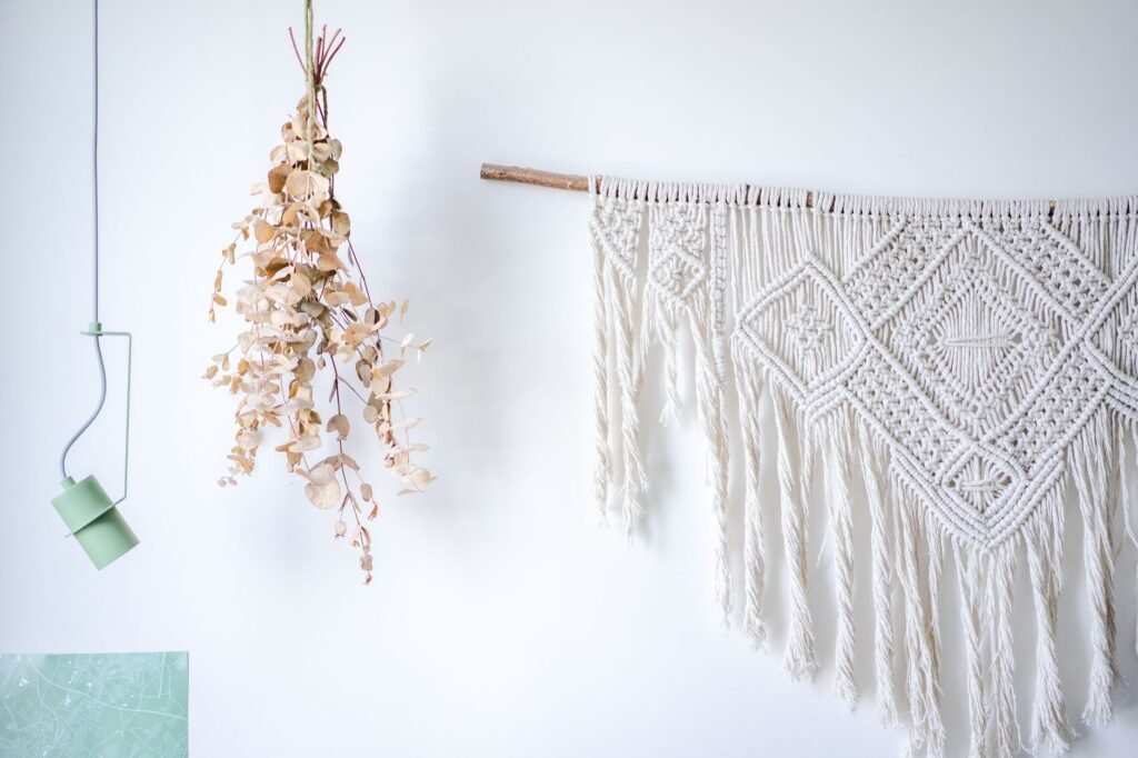 Cream macrame hanging with knotted diamond patterns and long fringe on a white wall beside dried flowers