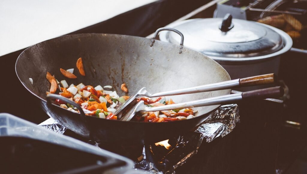 Chopped vegetables being stirred with metal spatulas in a large wok over a burner