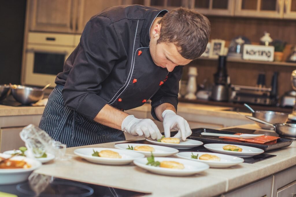 Chef in a dark jacket and gloves plating small portions onto white plates at a kitchen counter