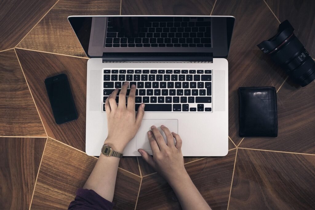 Hands typing on a silver laptop on a wooden desk beside a phone, wallet and camera