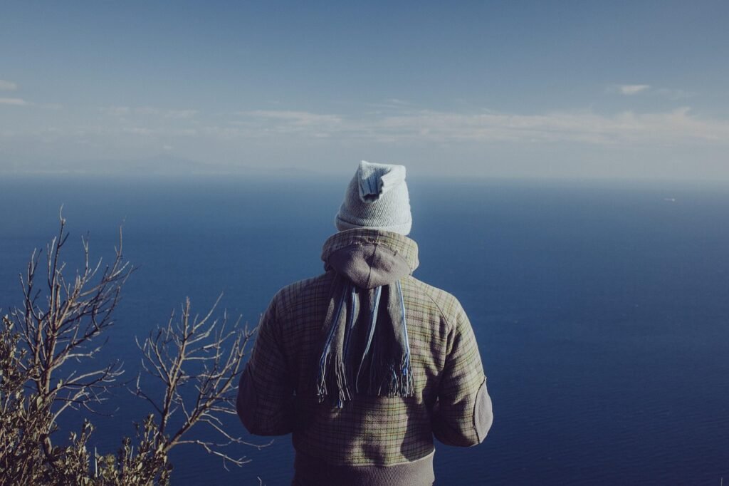 Person in a knit hat, hood and scarf looking out over a cold blue sea