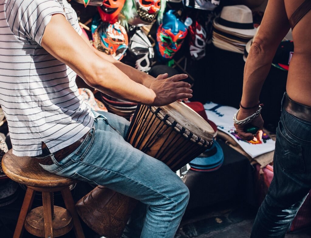 Man in a striped shirt playing a hand drum on a stool beside a market stall